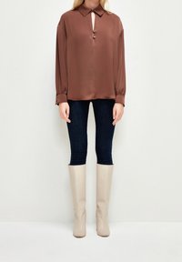 Brun satinbluse med krave og en nøglehulldetalje, parret med mørke denimjeans og knæhøje beige støvler. Glat tekstur, afslappet pasform.