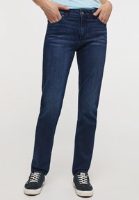 Osoba oblečená v tmavě modrých úzkých džínách a námořnických teniskách s bílými tkaničkami, s rukama položenýma v předních kapsách, na jednobarevném pozadí.