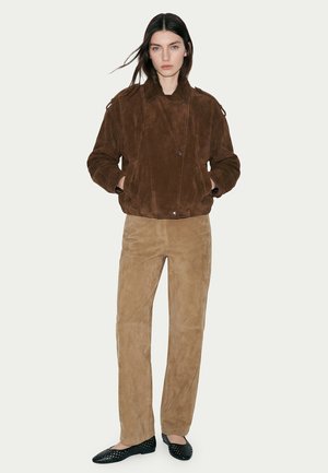 Donna con capelli lunghi e scuri che indossa una giacca in camoscio marrone, pantaloni in camoscio color cuoio e ballerine nere traforate, in piedi davanti a uno sfondo semplice.