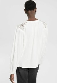 Camicia bianca a maniche lunghe con schiena plissettata e dettagli in pizzo floreale sulle spalle, con polsini aderenti e vestibilità comoda.