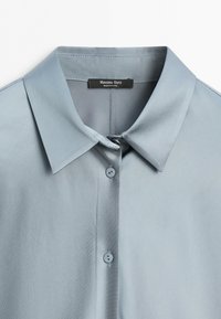 Camicia azzurro chiaro con collo, due bottoni visibili e un'etichetta nera con la scritta Massimo Dutti made in China all'interno dello scollo.
