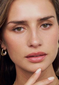 Nærbillede af et ansigt med olivenfarvet hud. Gyldne hoops øreringe og en subtil lipfarve. Glat struktur og naturlig makeup, med fingrene hvilede på hagen.