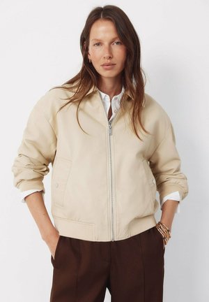 Frau mit langen braunen Haaren, die eine beige Jacke mit Reißverschluss über einem weißen Hemd und dunkelbraunen Hosen trägt, steht mit den Händen in den Taschen vor einem weißen Hintergrund.