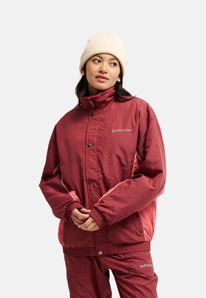 Junge Frau trägt eine cremefarbene Mütze und eine bordeauxrote Jacke mit passenden Hosen, steht mit verschränkten Händen und schaut zur Seite.