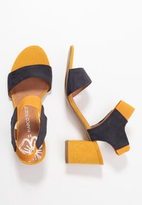 Avoimet kärjelliset korkokengät, joissa on mustasta mokasta valmistettu yläosa ja keltaiset mokasta valmistetut varvas- ja nilkkahihnat. Loiva korko, jolla on sileä pinta.