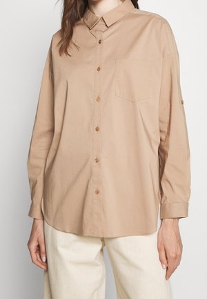 Kvinde iført en løs beige skjorte med brystlomme og cremefarvede bukser mod en ensfarvet baggrund.