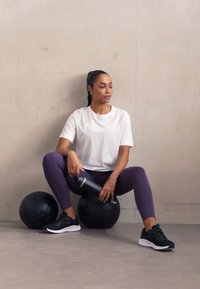 Juodos mankštos kamuoliai ant pilko grindų, o asmuo baltu marškinėliu ir violetinėmis Leggins sėdi, laikydamas sidabrinį vandens butelį.