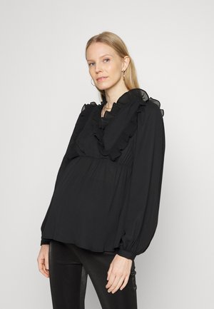 Blusa preta feita de tecido leve, com um decote com babados, detalhes plissados e mangas bufantes longas. Estilizada com calças pretas justas.