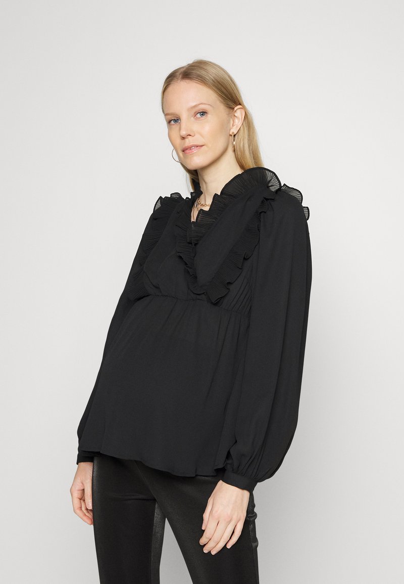 Blusa preta feita de tecido leve, com um decote com babados, detalhes plissados e mangas bufantes longas. Estilizada com calças pretas justas.