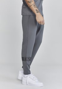 Pantalones atléticos grises hechos de una tela elástica, con una banda de logo negra en el tobillo y combinados con zapatillas blancas.