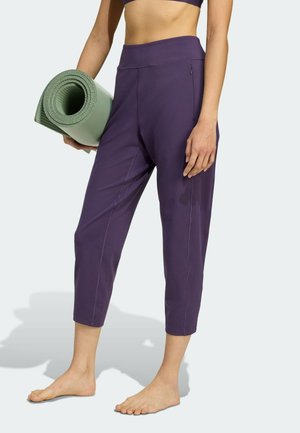 Frau in lila verkürzten Leggings, die eine zusammengerollte grüne Yogamatte hält und barfuß vor neutralem Hintergrund steht.