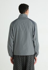 Veste légère grise avec un col haut, des poignets élastiques et des accents blancs. Présente des panneaux en maille sur les épaules pour une aération.