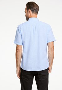 Vaalean sininen lyhythihainen paita, joka on valmistettu tekstuuripinnasta, jossa on klassinen kaulus ja laskosyksityiskohta takana, yhdistettynä mustiin housuihin.