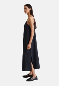 Schwarzes ärmelloses Kleid mit lockerem Schnitt, seitlichen Schlitzen, aus leichtem Stoff. Kombiniert mit schwarzen Schuhen mit einer Schnallenakzent.