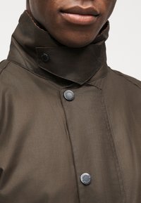 Κοντινό πλάνο του κάτω μέρους του προσώπου και του λαιμού ενός ατόμου που φοράει σκούρο καφέ μπουφάν με ψηλό γιακά και κουμπιά με το λογότυπο της μάρκας.