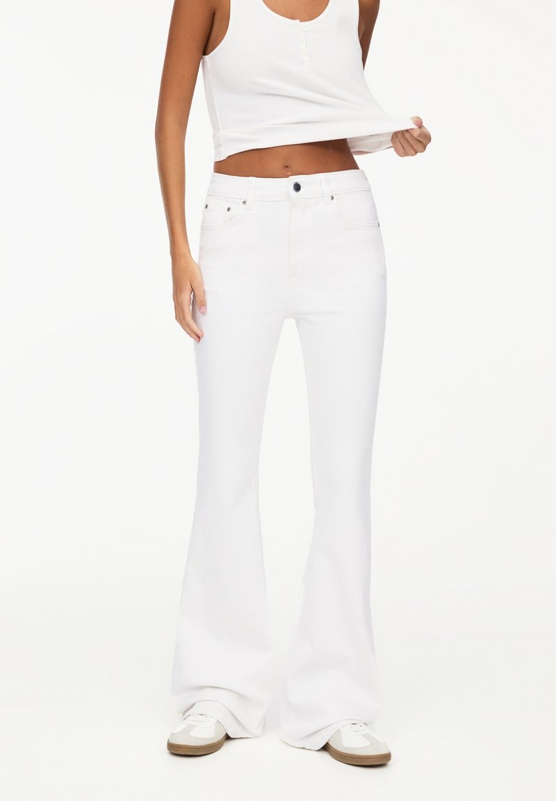 Personne portant un jean évasé blanc, des baskets blanches et un haut sans manches blanc, soulevant l’ourlet pour montrer la zone de la taille.