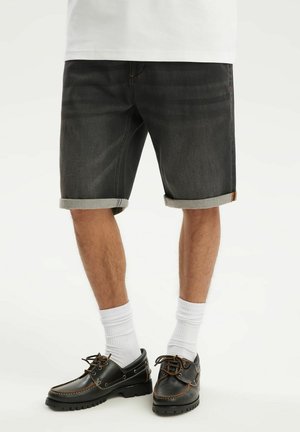 Mężczyzna w podwiniętych czarnych krótkich spodenkach jeansowych, białych skarpetach oraz czarnych skórzanych butach sznurowanych, stojący na białym tle.