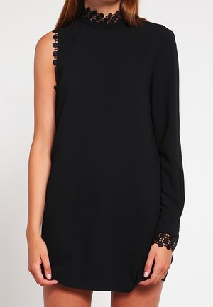 Kvinde iført en sort asymmetrisk kjole med én lang ærme og én ærmeløs side, med blonde detaljer på krave og håndled.