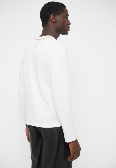 Biela dlhá blúzka s okrúhlym výstrihom a zlatým reťazovým detailom na chrbte. Materiál pôsobí mäkko a neformálne. Žiadne viditeľné vzory.