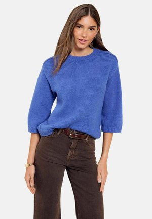 Femme portant un pull en tricot bleu à manches trois-quarts et un pantalon marron taille haute avec une ceinture marron, debout avec une main sur la hanche.