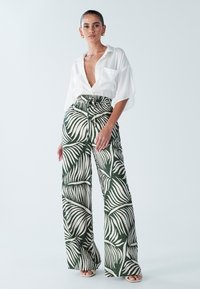 Hlače širokih hlačnic v zeleni in bež barvi z vzorcem palminega lista; kombinirane z ohlapno belo bluzo z globokim V-izrezom in kratkimi rokavi.