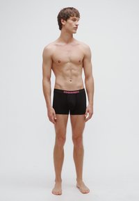 Fekete alsónemű texturált mintával, rózsaszín "DSQUARED2" derékpánttal. A modell mezítláb áll, előre nézve.