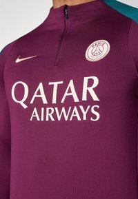 Fioletowa koszulka sportowa z pół zamkiem, z białym napisem "QATAR AIRWAYS" oraz okrągłym logo. Niebieskie akcenty na ramionach. Widoczne logo Nike.