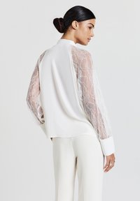 Blusa bianca con maniche in pizzo trasparente e retro in tessuto solido, dotata di colletto e polsini larghi. Tessuto liscio con dettagli in pizzo intricati.