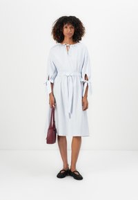 Mulher com cabelo encaracolado, vestindo um vestido midi riscado azul claro com mangas atadas, segurando uma carteira cor de vinho e usando sapatos planos pretos.