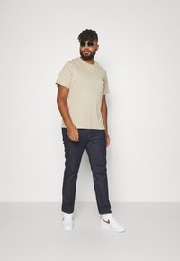 Tricou bej deschis cu un grafic mic pe piept, asociat cu blugi de un albastru închis și pantofi albi cu un accent maro. Ochelari de soare purtați.