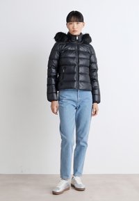 Giubbotto imbottito nero con cappuccio foderato in pelliccia, chiusura con zip e vestibilità oversized. Abbinato a jeans azzurri chiari e sneakers argentate.
