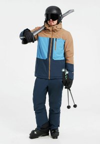 Conjunto de esquí que incluye una chaqueta marrón y azul, pantalones azul marino, casco negro y guantes. Los esquís y los bastones se sostienen sobre el hombro.
