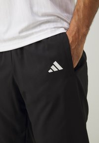 Black athletic pants with a smooth texture, featuring a white Adidas logo on the left thigh and a relaxed fit design.