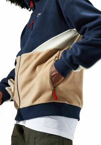 Fleecejacke mit Farbbereichen in Marineblau, Creme und Beige. Verfügt über Reißverschlusstaschen und gerippte Bündchen. Strukturierter, warmer Stoff für zusätzlichen Komfort.