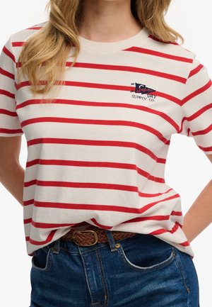 Vrouw draagt een wit T-shirt met rode horizontale strepen en een klein marineblauw en rood logo op de borst, gecombineerd met een blauwe spijkerbroek en een bruine gevlochten riem.