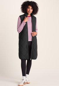 Woman wearing a long black quilted vest over a red and white striped long-sleeve shirt, black leggings, white socks, and white sneakers.