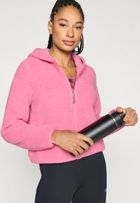 Różowa, miękka bluza z kapturem na zamek, wykonana z teksturowanego materiału. W jednej ręce trzymany jest czarny termos, w parze z czarnymi legginsami.