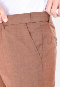 Pantaloni marroni, strutturati, con taglio dritto; vita e tasche laterali visibili. Il tessuto presenta un sottile motivo intrecciato.
