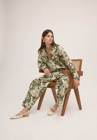 Femme assise sur une chaise en bois, portant une blouse et un pantalon à motifs floraux assortis avec des ballerines couleur crème, sur fond neutre.