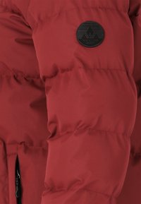Rød pufferjakke med quiltet design, der har en rund sort logo-patch på ærmet og en lynlåslomme på siden. Glat tekstur.