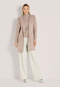 Frau in beigem Strickcardigan und Pullover mit weißen, weit geschnittenen Hosen und silbernen spitzen Schuhen, die vor einem schlichten Hintergrund steht.