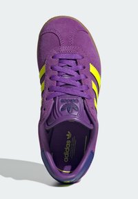 Fioletowy zamszowy sneaker z żółtymi paskami, teksturowanymi akcentami i sznurowanym designem. Posiada gumową podeszwę oraz logo Adidas na języku i wkładce.