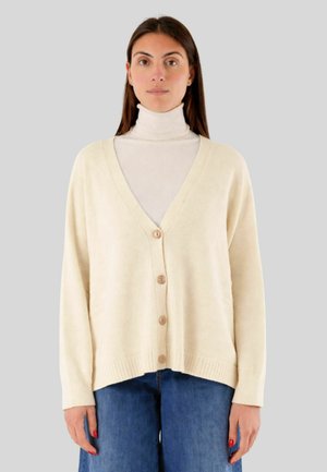 Donna che indossa un cardigan color crema con scollo a V e bottoni sopra un dolcevita bianco e jeans blu, in piedi davanti a uno sfondo grigio.
