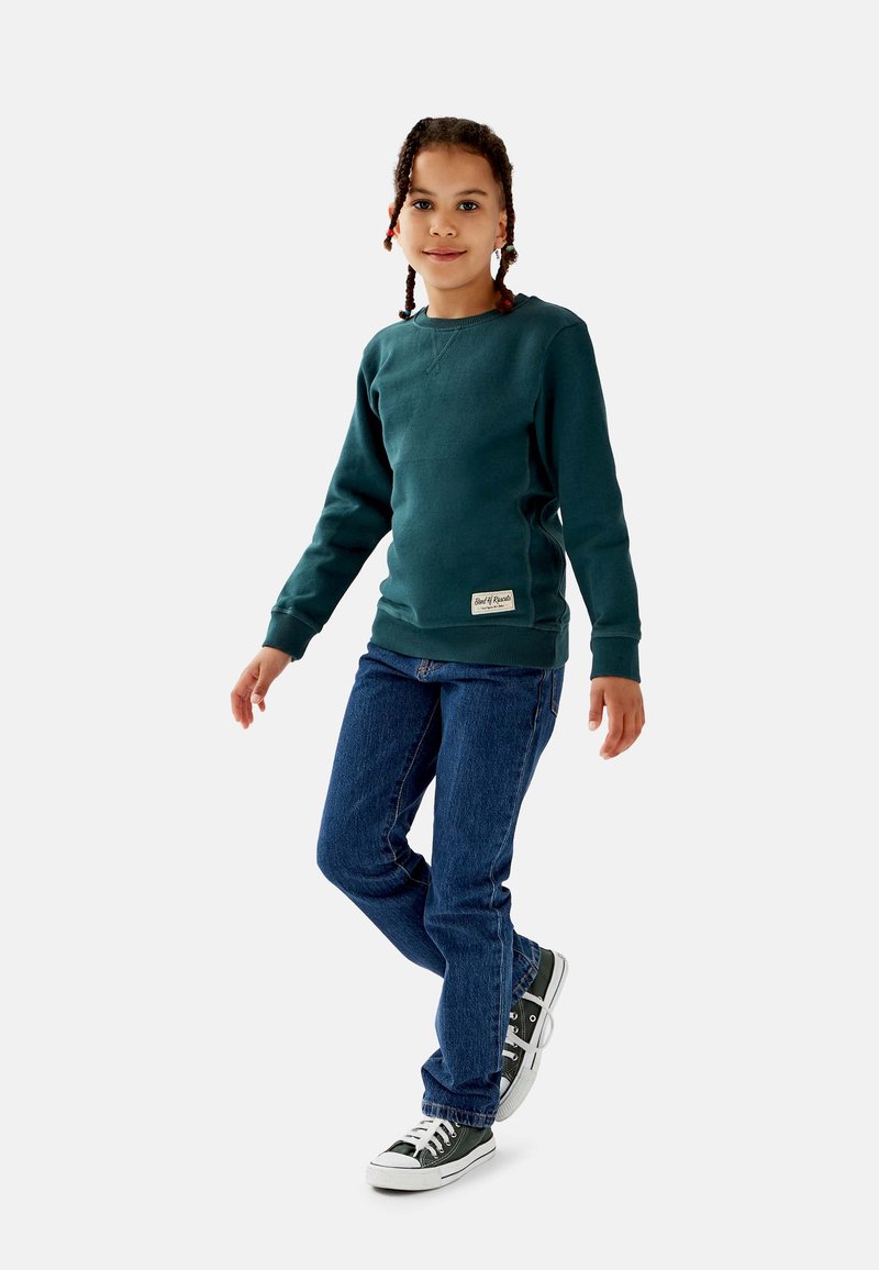Barn med fletninger har en teal sweatshirt, blå jeans og sorte sneakers på, står og vender fronten mod kameraet mod en ensfarvet hvid baggrund.