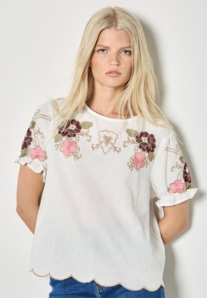 Junge Frau mit langen blonden Haaren, die eine weiße Bluse mit pinker und violetter Blumenstickerei und gewelltem Saum trägt, vor einem schlichten Hintergrund stehend.