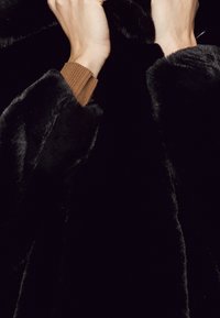 Sort pelsjakke i imiteret pels med en blød, glansfuld tekstur. Hænder holder om kraven; en brun ribbet manchet er delvist synlig.