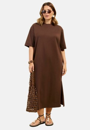 Kvinde iført brun midi kjole med sideslid, brun net-tote taske, mørke solbriller, skalsandaler og perlearmbånd, stående mod en hvid baggrund.