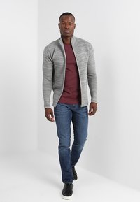 Graue, strukturierte Zip-Jacke über einem kastanienbraunen Shirt, kombiniert mit blauen Jeans und schwarzen Sneakers. Das Model steht vor einem hellen Hintergrund.