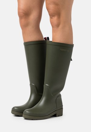 Jalat, joissa on polven korkeudelle ulottuvat oliivinvihreät kumisadetossut, joilla on uritetut pohjat ja pieni vetolenkki takana, yksinkertaisella taustalla.