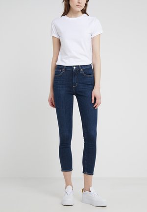 Kobieta ubrana w białą koszulkę z krótkim rękawem, ciemnoniebieskie, wysokie spodnie skinny skrócone powyżej kostek oraz białe sneakersy na platformie.